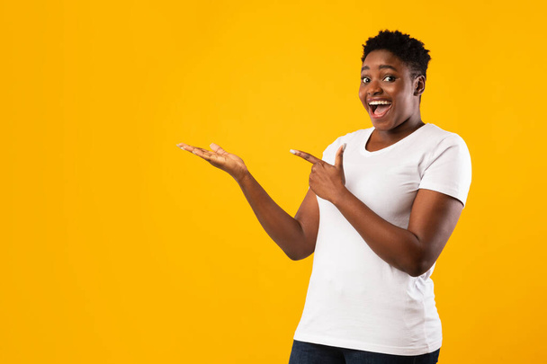 Joyful Plus-Size africano signora che indica dito che tiene qualcosa, sfondo giallo - Foto, immagini