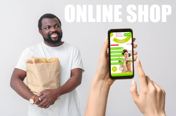 Nainen, jolla on matkapuhelin, jossa on avoin sivu elintarvikkeiden jakelupalvelua ja afroamerikkalainen mies, jolla on tuoreita tuotteita markkinoilta harmaalla taustalla - Valokuva, kuva