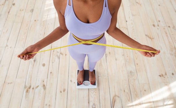 Close-up de mulher negra magro medindo sua cintura enquanto em pé em escalas em casa, vista acima - Foto, Imagem