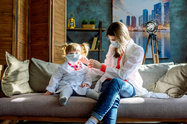 Two cute little girls sisters are playing doctor at home on the couch. Doctor uniform, respirator mask, protection against coronavirus. COVID-19 - Foto, Imagem
