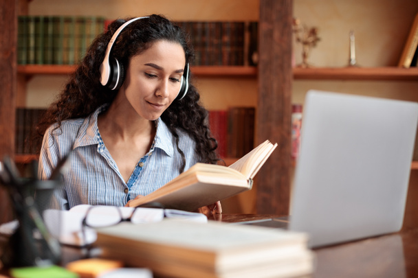 Lady sitting at desk, wearing headset, using computer, reading book - Photo, Image