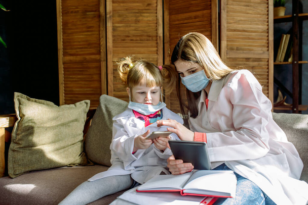 Two cute children play doctor and hospital using stethoscope. girl in uniform of a doctor and in face mask. Coronavirus concept, covid-19.Friends girls having fun at home or preschool. - Foto, Imagen