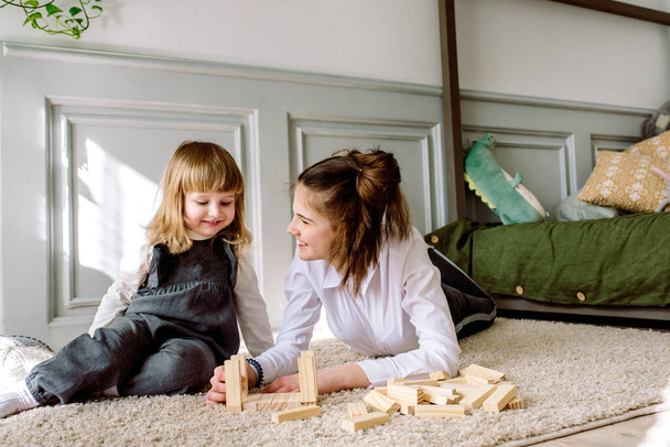 two cheerful cute little girls sisters play on the floor with wooden blocks. Cozy family games at home.concept of family and child.Bright sunny room,bed and exotic indoor plants - Photo, Image