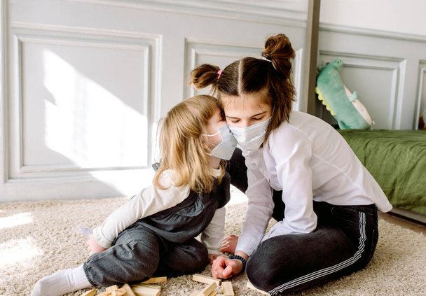 two cheerful little girls sisters play on floor with wooden blocks. Cozy family games at home.On carnine at home, coronavirus covid-19.Interaction and communication of children in face mask. - Photo, image