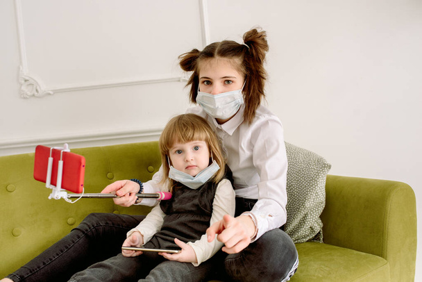 Two girls play a doctor at home in a bright room. On a green sofa they play with a bear, bandage a foot to a toy. In a face mask, protection against Chovid-19 virus.Big sister takes selfie for blog - Photo, Image
