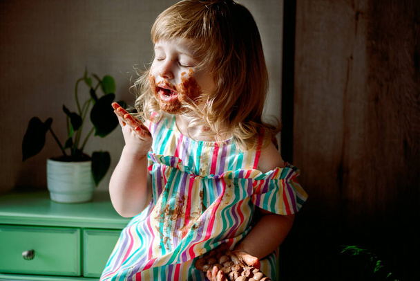 Cute curly haired baby girl eating chocolate,child smeared. dirty cheeks, nose and hands. Chocolate celebration on July 7th. harmful sweets for children. unhealthy food. indoor plants. - Photo, Image