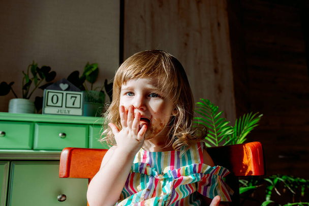 Cute curly baby girl eating chocolate. The child is allergic to the sweet, dirty girl, July 7 is chocolate day. Homes, indoor plants. - Zdjęcie, obraz