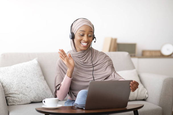 Szczęśliwy czarny muzułmanin lady w hidżabie i słuchawki podejmowania rozmowy wideo na laptopie w domu, machając na ekranie - Zdjęcie, obraz