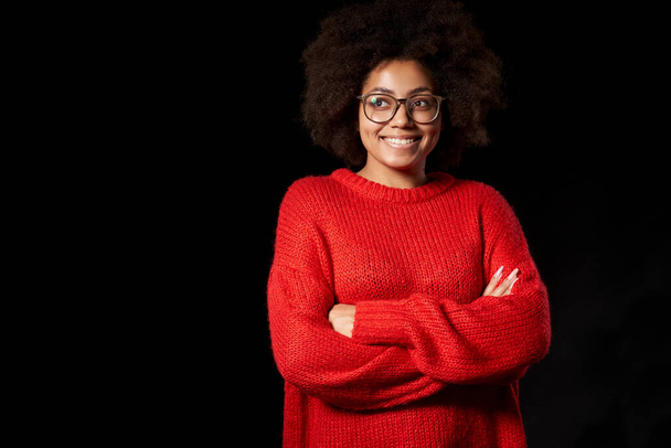 Happy mixed race girl in glasses and red sweater, standing with folded hands, looking to side - Фото, зображення