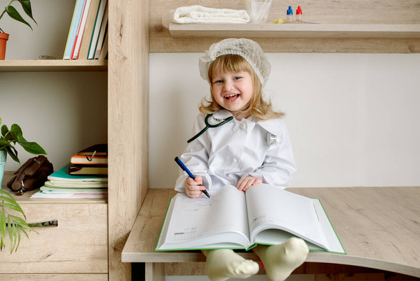 baby girl uniformed as doctor writing to clipboard.Medical gown and phonendoscope. The doctor in the ward writes a medical history - Фото, изображение