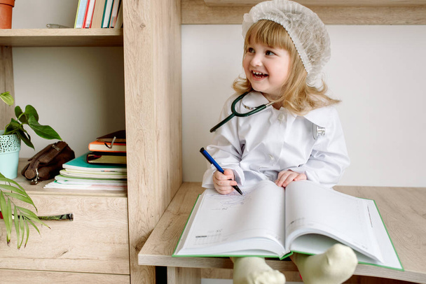 Cute little baby girl in doctor uniform with clipboard in hospital.Phonendoscope and white bathrobe. Notepad and history of disease. The concept of health, medicine and laboratory diagnostics. - 写真・画像