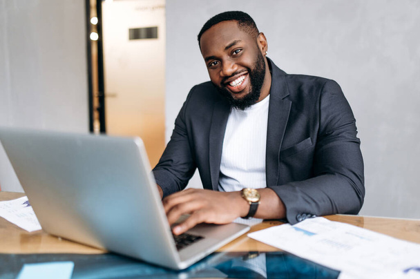 Portret afrykańskiego biznesmena w miejscu pracy. Udany menedżer płci męskiej przy użyciu laptopa, pracy nad nowym projektem lub startupem. Przystojny, pewny siebie pracownik patrzy w obiektyw, uśmiechając się - Zdjęcie, obraz