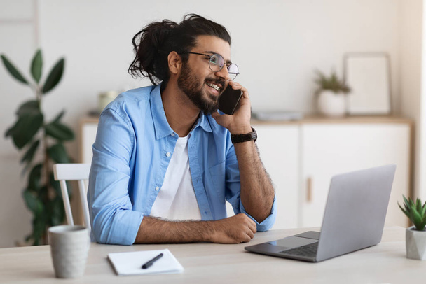 Fiatal arab férfi beszél mobilon, dolgozik a laptop otthoni irodában - Fotó, kép