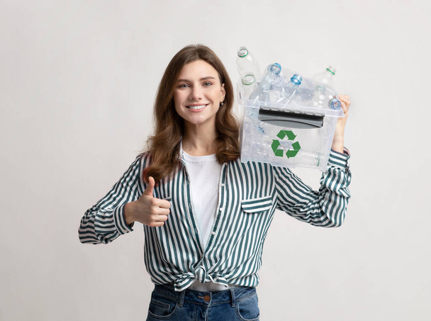 Lächelnde Frau hält Papierkorb mit Plastikflaschen und zeigt Daumen hoch - Foto, Bild