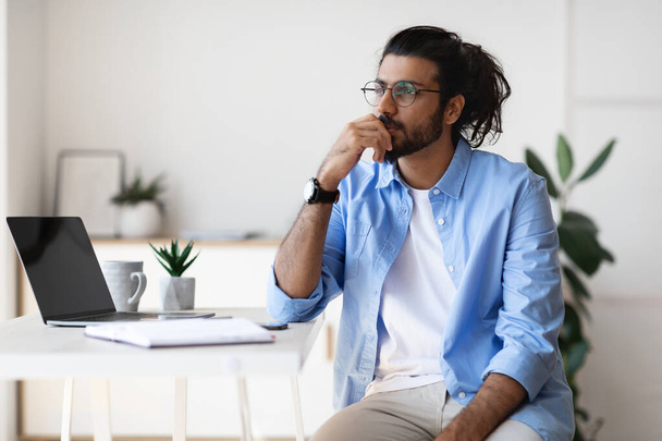 Thoughtful young arab male freelancer sitting at desk at home, looking aside - Фото, изображение