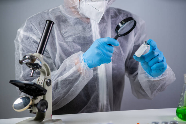 Healthcare and medical concept. Pharmaceutical research and clinical trials concept. Laborer in protective clothing dispenser and glasses is examining new medicine in ampoules under magnifying glass. - Photo, Image