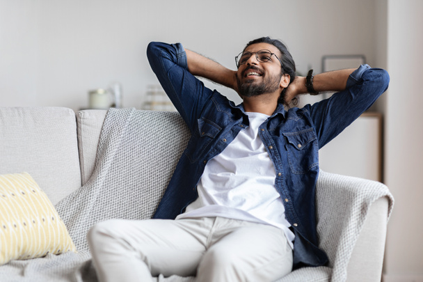 Расслабленный улыбающийся араб, откидывающийся на диване, расслабляющийся дома - Фото, изображение