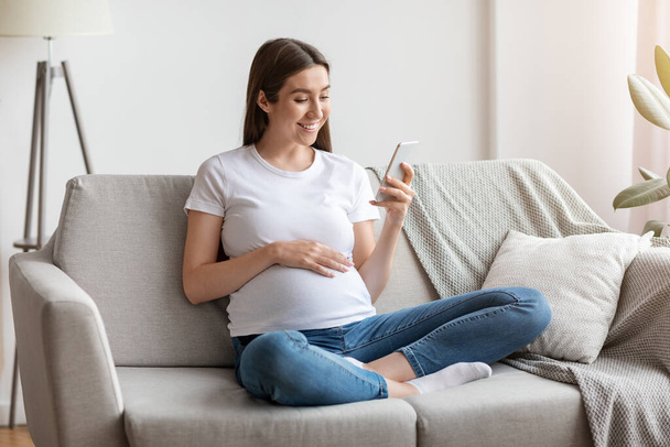Счастливая беременная женщина пользуется смартфоном дома, просматривая приложение о беременности - Фото, изображение