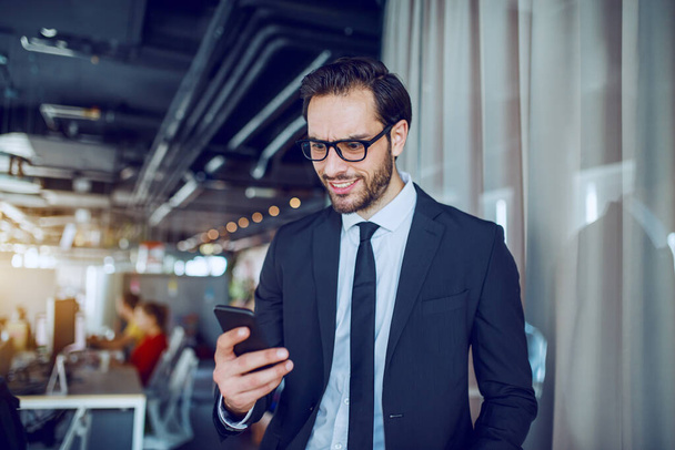Attractive caucasian businessman in suit and with eyeglasses standing in company and reading message on smart phone. In background are employees working. - Foto, imagen