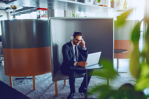 Empresário barbudo caucasiano alegre em terno, com óculos e fones de ouvido sentados no local de trabalho e usando laptop. - Foto, Imagem