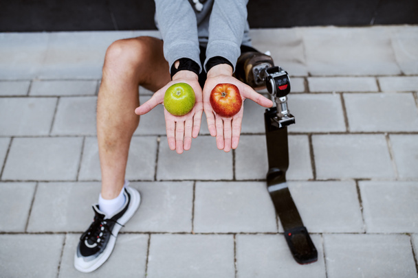 Vue de dessus de l'homme avec la jambe artificielle assise sur le sol et tenant des pommes rouges et vertes dans chaque main. - Photo, image