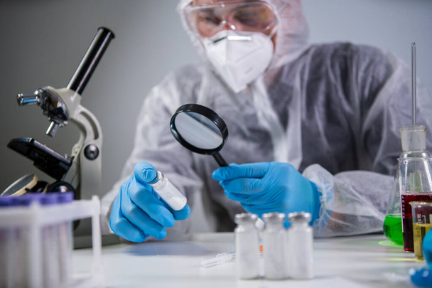 Healthcare and medical concept. Pharmaceutical research and clinical trials concept. Laborer in protective clothing dispenser and glasses is examining new medicine in ampoules under magnifying glass. - Photo, Image