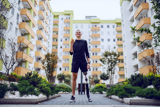Ganzer Länge attraktiver kaukasischer Sportler mit künstlichem Bein, der im Park steht, umgeben von Gebäuden und Bananen isst. - Foto, Bild