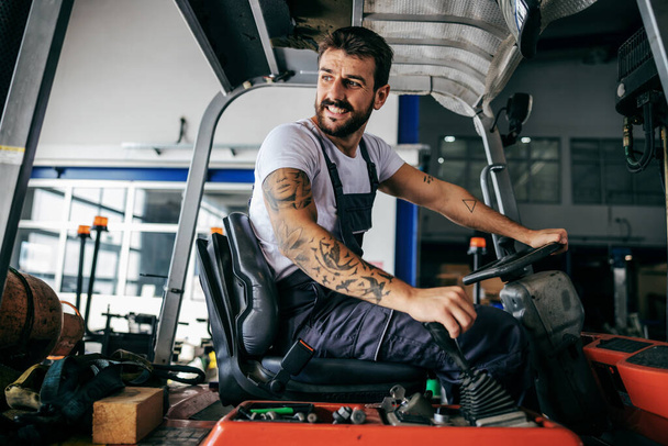 Smiling bearded tattooed hardworking employee in overalls sitting in vehicle and driving it. - Fotoğraf, Görsel