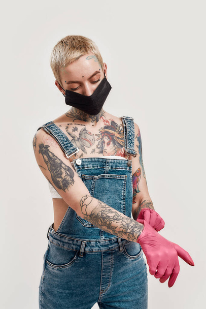 Quarantine accessories. A white tattooed and pierced woman wearing black respiratory mask with rubber gloves and denim overalls - Foto, afbeelding