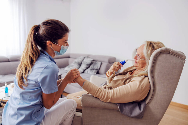 Idősebb nő ül otthon a székben, és lélegzi be az oxigént a lélegeztetőgépből. A nővér mellette ült, fogta a kezét és vigasztalta.. - Fotó, kép