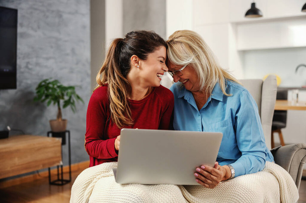 Χαμογελώντας μητέρα και κόρη κάθονται στο σπίτι μαζί και χρησιμοποιούν φορητό υπολογιστή για να κρέμονται στα μέσα κοινωνικής δικτύωσης. - Φωτογραφία, εικόνα