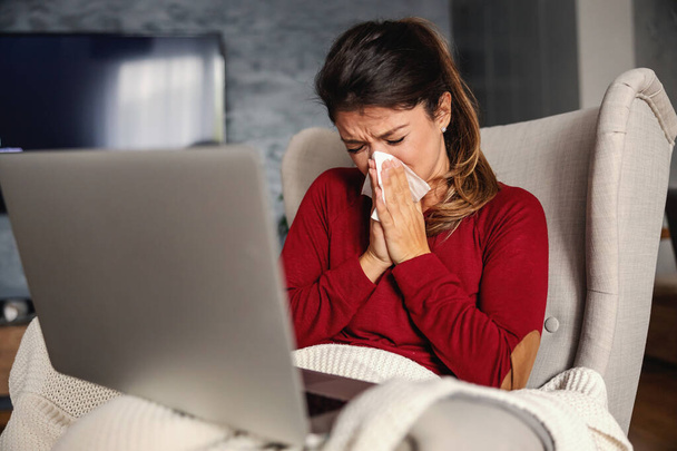 Femme malade assise dans une chaise à la maison pendant le confinement avec un ordinateur portable dans ses genoux et le nez soufflant. - Photo, image