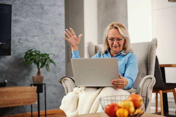 Mosolygó, izgatott, idős nő ül a székben otthon és laptopot néz. A közösségi médián lóg.. - Fotó, kép