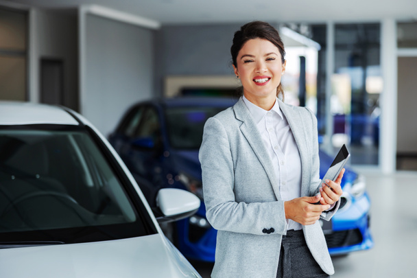 Barátságos, mosolygós női autó eladó áll autószalon és gazdaság tabletta, miközben a kamera. - Fotó, kép