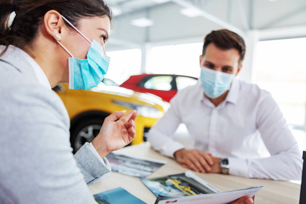 Car seller and customer sitting at the table in car salon and talking about specifications and car performances during corona virus. They both have face masks on. - Фото, зображення