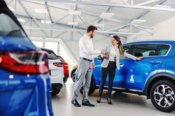 Amistoso, sonriente vendedor femenino mostrando coche nuevo a un cliente mientras está de pie en el salón de coches. - Foto, imagen