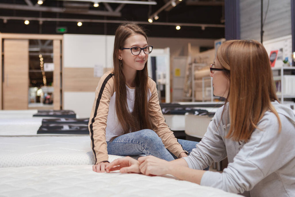 Happy teen girl talking to her mom while choosing new orthopedic mattress to buy - Foto, afbeelding