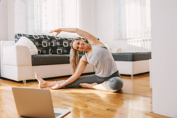 Сидя дома на полу и показывая упражнения на растяжку. У неё онлайн-занятия за ноутбуком.. - Фото, изображение