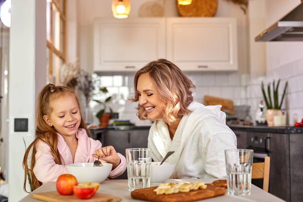 happy caucasian adult woman and child girl have breakfast in the kitchen at home - Φωτογραφία, εικόνα