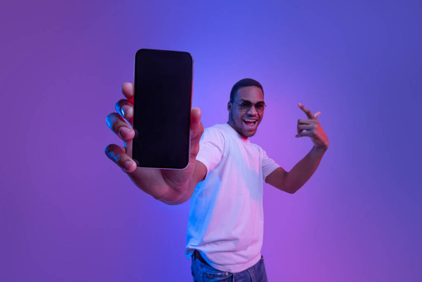 Oferta móvil. Emocionado chico africano demostrando Smartphone con pantalla negra, Iluminación de neón - Foto, imagen