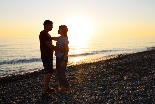 Satisfied man and woman stand close, hug and look at each other at sunset on the sea beach. Happy relationship concept for Valentine's day - Photo, Image