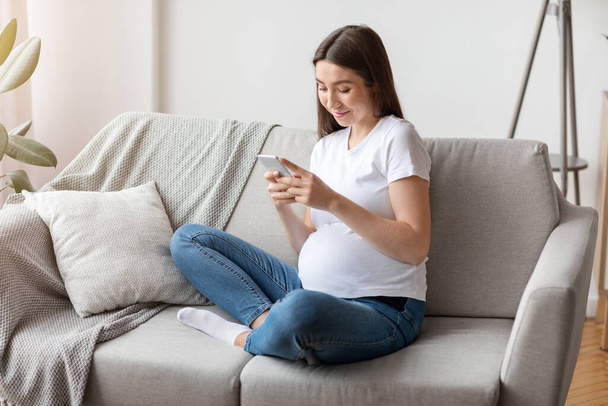 Беременность Tracker App. Улыбающаяся молодая беременная женщина с помощью смартфона дома - Фото, изображение