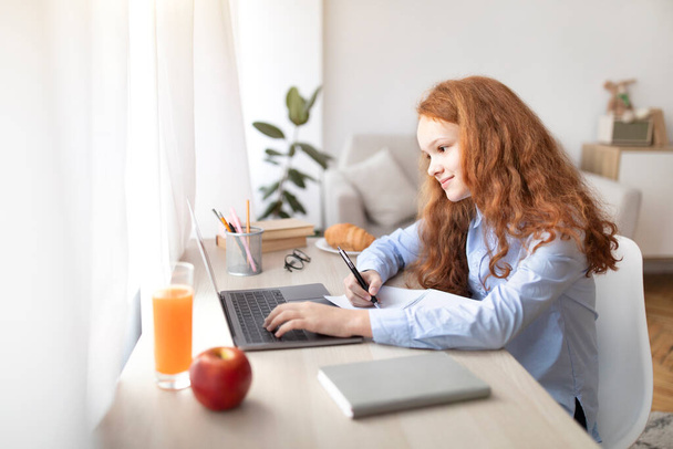Egy lány ül az asztalnál, laptopot használ, és a füzetébe ír. - Fotó, kép
