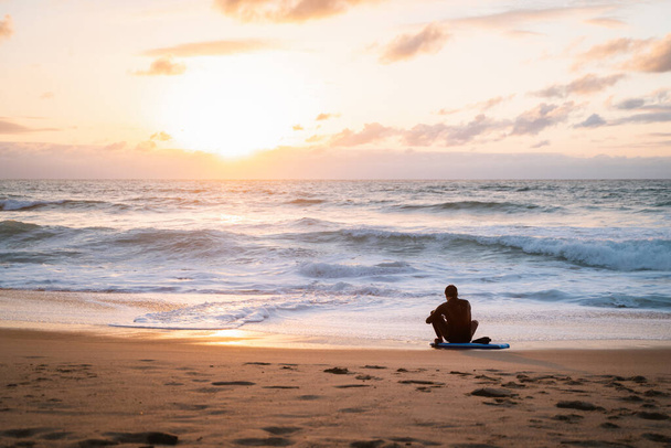 Молодий чоловік сидить на дошці свого тіла на березі самотнього пляжу під час заходу сонця коригуючи свій мокрий костюм
 - Фото, зображення