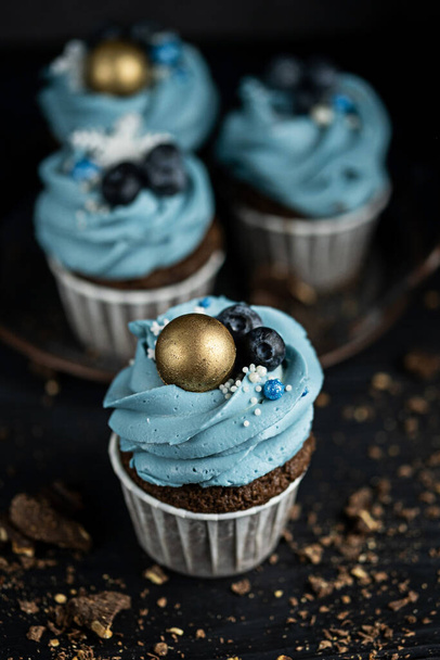 Several muffins or cupcakes with blue shaped cream and blueberrieson at black table. Rustic style copyspace. - Фото, зображення