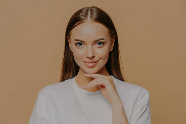 Lovely young European woman with makeup healthy skin touches gently jawline looks directly at camera has long straigh hair natural beauty dressed in casual sweater isolated over brown background - Photo, Image