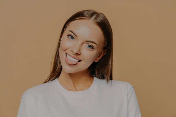 Portrait of playful dark haired woman tilts head shows tongue and smiles positively has fun indoor dressed in white sweater poses against beige background. Human face expressions and positive feelings - Photo, Image