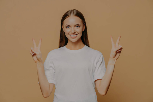 Horizontal shot of happy optimistic young Caucasian woman raises arms shows peace gesture with fingers does victory sign says number two wears casual white t shirt isolated over brown background - Photo, Image