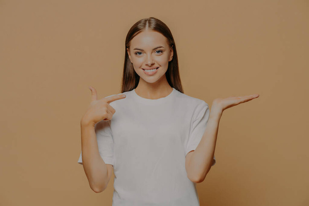 Half length shot of good looking brunette young woman points at raised palm pretends holding something advertises some item wears casual t shirt isolated over brown background. Promotion concept - Photo, Image