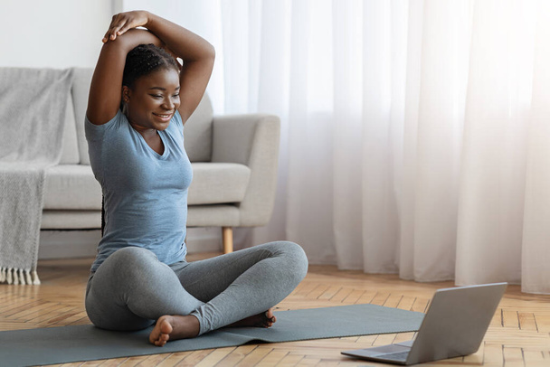 Sportliche schwarze Dame dehnt sich vor Laptop zu Hause, Online-Training - Foto, Bild